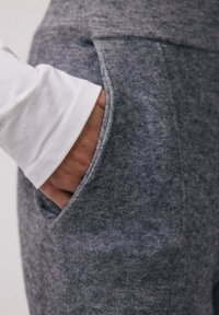 Main dans la poche d'un pantalon en polaire gris porté avec une chemise blanche à manches longues, montrant les détails de texture et de couture.