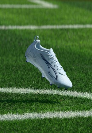 Clapă albă de fotbal cu design negru suspendată deasupra unui teren cu iarbă verde, lângă linii de delimitare albe.