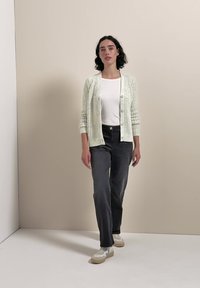 Donna in piedi con cardigan bianco strutturato sopra top bianco, jeans neri e sneakers bianche contro sfondo di parete beige.