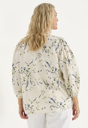 Vrouw met blond golvend haar, gekleed in een crèmekleurige blouse met blauwe bloemenprints en volumineuze mouwen, staand tegen een effen achtergrond.
