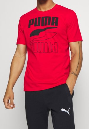 Persoon draagt een rood Puma T-shirt met zwart logo en zwarte Puma broek, staand met één hand in de zak tegen een effen achtergrond.