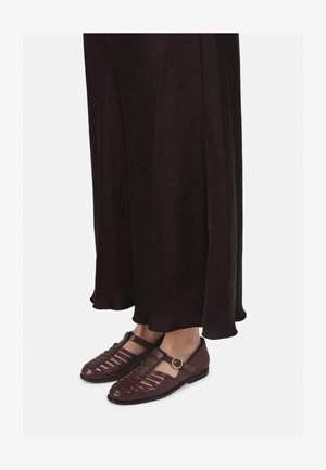 Brūni ādas apavi ar atvērtu austuvju dizainu un sānu sprādzi. Apģērbs sastāv no garas melnas plūstošas svārku.