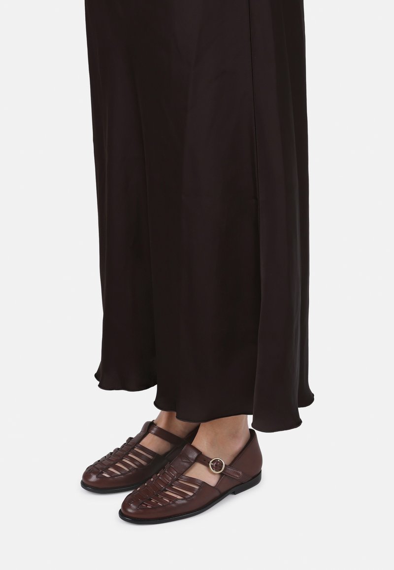 Scarpe in pelle marrone con design intrecciato aperto e una fibbia laterale. L'outfit è composto da una lunga gonna nera e fluida.