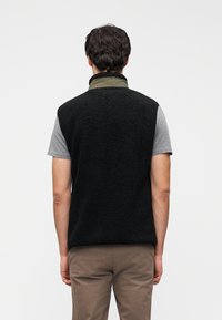 Μαύρο αμάνικο γιλέκο από φλις με ψηλό γιακά, σε συνδυασμό με γκρι κοντομάνικο πουκάμισο από κάτω. Υφή μαλακού, αφράτου υλικού· casual σχεδιασμός.