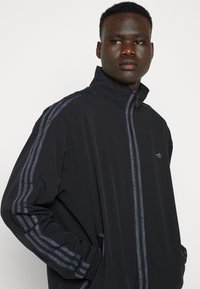 Veste de sport noire avec un col haut. Présente des accents rayés bleus le long des manches et un logo subtil sur la poitrine. Poches zippées.