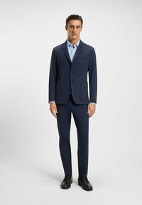 Tummansiniset räätälöidyt puvut, joissa on sileä pinta, yksirivinen takki ja hammaslapeet, yhdistettynä vaaleansiniseen ruutupaitaan ja mustiin kenkiin.