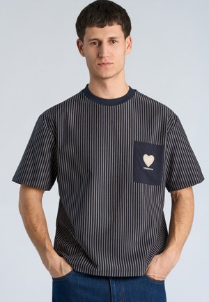 Junger Mann mit kurzen dunklen Haaren, der ein schwarz-weiß gestreiftes T-Shirt mit einer navyblauen Tasche mit Herzdesign und Text trägt.