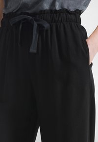 Svarta byxor med dragsko, elastisk midja, sidofickor och en mjuk, slät textur. Designen är avslappnad och casual.