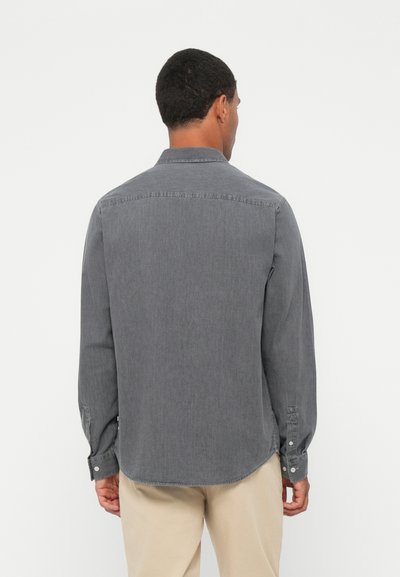 Chemise grise à manches longues en tissu doux, dotée d'un col pointu, de poignets boutonnés et d'un ourlet arrondi, portée avec un pantalon de couleur claire.