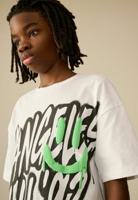 Weißes Baumwoll-T-Shirt mit auffälligem schwarzen Graffiti-Text und einem tropfenden grünen Smiley-Design, kurzen Ärmeln und Rundhalsausschnitt.