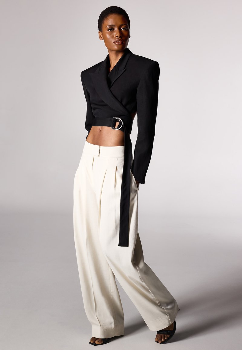 Blazer noir court avec un détail de ceinture argentée, associé à un pantalon large blanc, présentant une taille haute et des plis à l'avant. Textures lisses.
