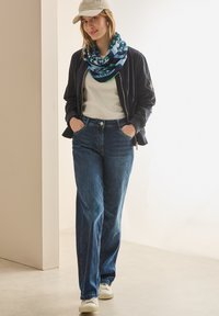 Afslappet outfit bestående af en navy bomberjakke, hvid t-shirt, blå jeans og et mønstret blå-grønt infinity tørklæde. Hvide sneakers fuldender looket.