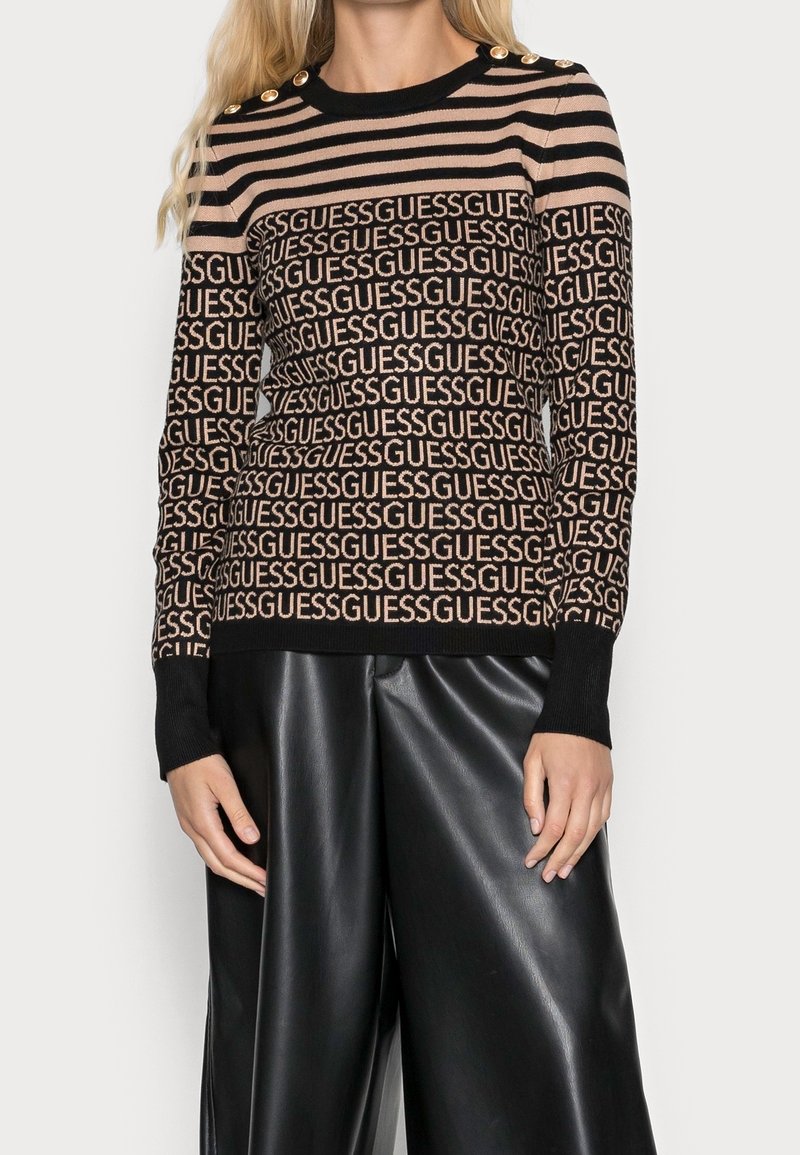Femme portant un pull rayé noir et beige avec le texte répété "GUESS" et des boutons dorés à l'épaule, associé à un pantalon large en cuir noir.