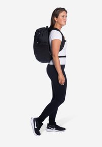 Vrouw in zwarte leggings, witte t-shirt, zwarte sneakers en een grote zwarte rugzak, staande in profiel tegen een witte achtergrond.