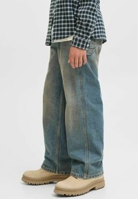 Svijetloplave široke traperice s izblijedjelim finišom i vidljivim šavovima. Uparen s bež gležnjačama. Iznad se nosi košulja s kariranim uzorkom.