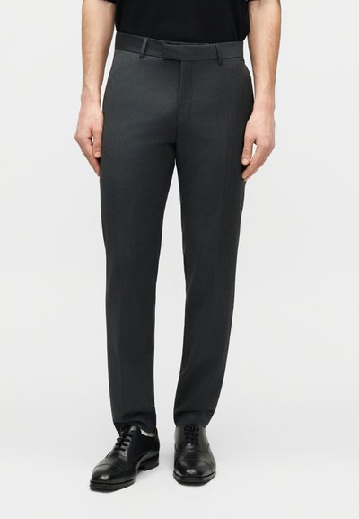 Άνδρας που φοράει σκούρο γκρι παντελόνι φόρμας και μαύρα δερμάτινα μοκασίνια, στέκεται μπροστά σε απλό φωτεινό φόντο.