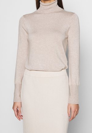 Donna con indosso un maglione beige a collo alto e una gonna midi crema a coste a vita alta, in piedi davanti a uno sfondo semplice.