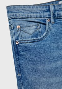 Gros plan sur un jean en denim bleu montrant la ceinture avant et la petite poche à monnaie avec des rivets en cuivre et des coutures visibles.