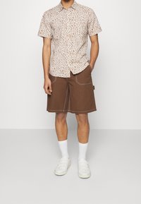 Camisa de manga corta en color crema claro con un patrón floral marrón; pantalones cortos marrones con costuras blancas; calcetines y zapatos blancos.