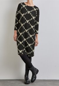 Robe noire avec un motif géométrique crème clair, manches trois-quarts, coupe ample, décolleté légèrement arrondi, portée avec des bottines noires.