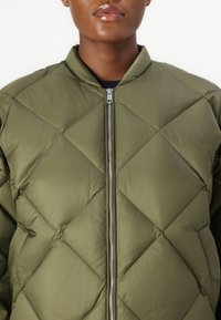 Veste matelassée olive vert avec fermeture éclair, présentant des motifs en losange et un col montant. Texture lisse et brillante.