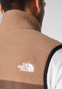 Smešani sme�đi flis prsluk s visokim ovratnikom, crnim rubom i bijelim izvezenim logotipom "The North Face" na gornjem dijelu leđa.