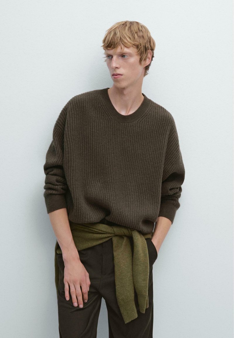 Junger Mann in einem dunkelbraunen Ripp-Pullover mit einem olivgrünen Pullover um die Taille, der vor einem einfarbigen hellen Hintergrund steht.
