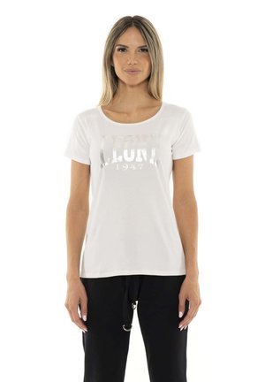 Une femme porte un T-shirt blanc à manches courtes avec un logo argenté et un pantalon noir sur un fond blanc uni.