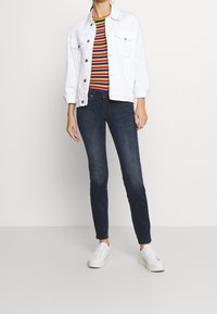 Chaqueta de mezclilla blanca sobre una camisa de rayas multicolores, combinada con jeans ajustados oscuros y zapatillas blancas con suelas texturizadas.