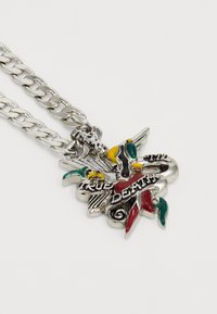 Colar de corrente prateada com um pendente colorido apresentando o texto "MORTE VERDADEIRA", com detalhes intrincados e acentos decorativos em vermelho, verde e amarelo.