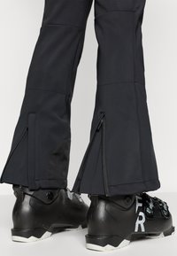 Czarne spodnie narciarskie z zamkami błyskawicznymi na mankietach, połączone z czarnymi butami narciarskimi z białymi akcentami i trzema zapięciami na klamry.