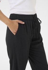 Černé, lehké kalhoty s elastickým pasem a šňůrkou. Obsahují boční kapsy a hladký povrch, zobrazené v úrovni boků.