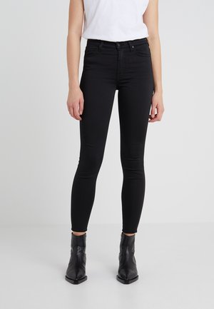 Personne portant un jean noir ajusté, des bottines noires et un haut blanc sans manches, debout devant un fond clair uni.
