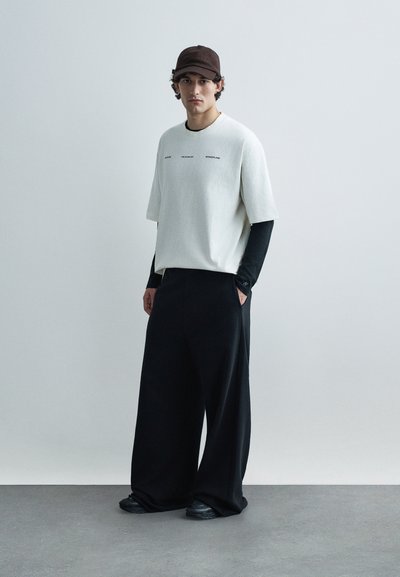 Biela košeľa s krátkym rukávom s čiernymi akcentmi, čierna vrstva s dlhým rukávom pod ňou, široké čierne nohavice a hnedá čiapka. Jednoduchý dizajn, voľný strih.