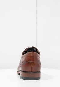 Chaussure habillée en cuir marron avec des détails en brogue, bout arrondi et talon empilé. Texture lisse et design élégant, vue de l'arrière.