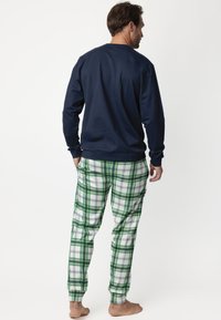 Hombre de pie descalzo, vistiendo una camisa de manga larga azul marino y pantalones blanquiverdes a cuadros, de espaldas contra un fondo blanco.