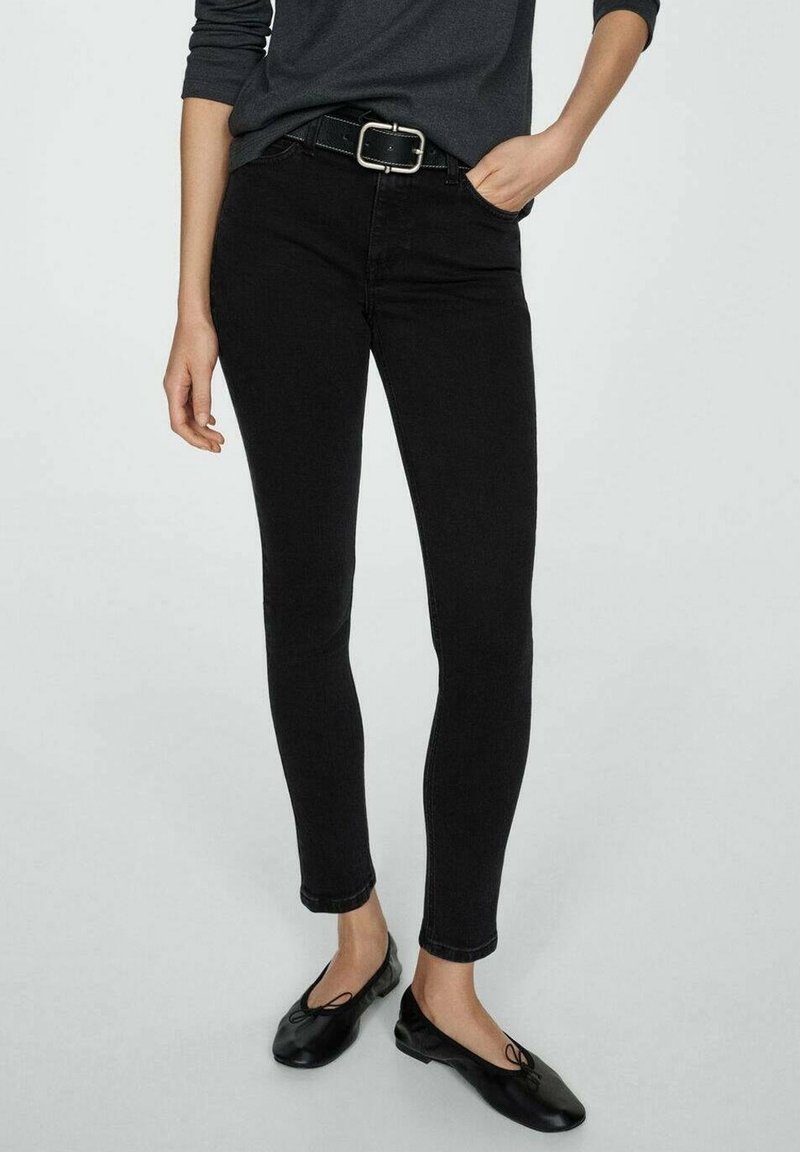 Femme portant un jean noir moulant, une ceinture noire, un haut sombre à manches longues et des ballerines noires, debout avec une main dans la poche.