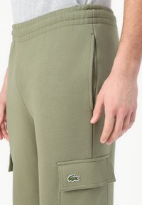 Olivově zelené cargo kalhoty vyrobené z měkké tkaniny, s bočními kapsami a vyšitým logem na stehně. Elastický pas pro pohodlí.