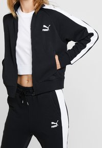 Svart zip-up jacka med vita sidostrick och Puma-logotyp; matchad med svarta joggers med sidostrick och logotyp. Slät tygtextur.
