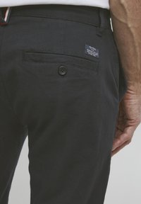 Melni bikses, izgatavotas no teksturēta auduma, ar aizmugures kabatu ar pogu un auduma etiķeti. Dizains ir vienkāršs un klasisks.