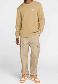 Man som bär en beige Nike-tröja, beige cargobyxor och vita och orange Nike-sneakers som står mot en enkel bakgrund.