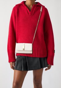 Punainen neulepusero, jossa on raidoitettu helma ja kaulus, yhdistetty valkoiseen ketjunauhakankaiseen käsilaukkuun. Musta pleated-ruudullinen hame.