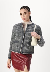 Pelēks adīts kardigans ar teksturētu rakstu, apaļu kakla izgriezumu un divām priekšējām kabatām, saskaņots ar bordo ādas mini svārkiem.