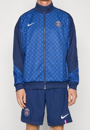 Man draagt een blauw Paris Saint-Germain sportjack met geometrisch patroon en bijpassende korte broek met team- en Nike-logo's.