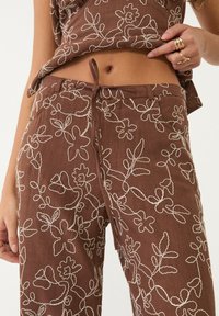 Brązowe lniane spodnie z białym haftem kwiatowym, z elastycznym pasem i subtelnymi detalami kieszeni. Teksturowana tkanina w luźnym fasonie.
