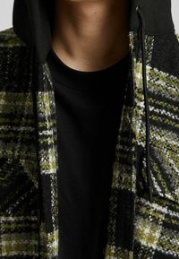 Ulljacka i grön och svart rutigt mönster med svart huva. Har en framzipper och dragsko detaljer, som visar en mjuk textur.