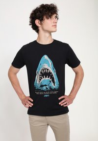 Czarny bawełniany t-shirt z grafiką niebieskiej rekina z otwartymi szczękami, z białym tekstem poniżej, przedstawiający scenę z "Szczęk".