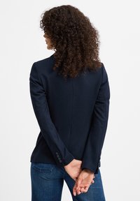 Marineblauer taillierter Blazer mit strukturierten Schultern, einzelner Rückenbelüftung und geknöpften Manschetten, kombiniert mit blauen Jeans. Glatte Stofftextur.