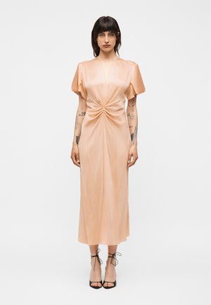 Kobieta o krótkich ciemnych włosach, ubrana w brzoskwiniową sukienkę midi z satyny z marszczonym przodem i bufkami, stojąca z widocznymi tatuażami na ramionach.