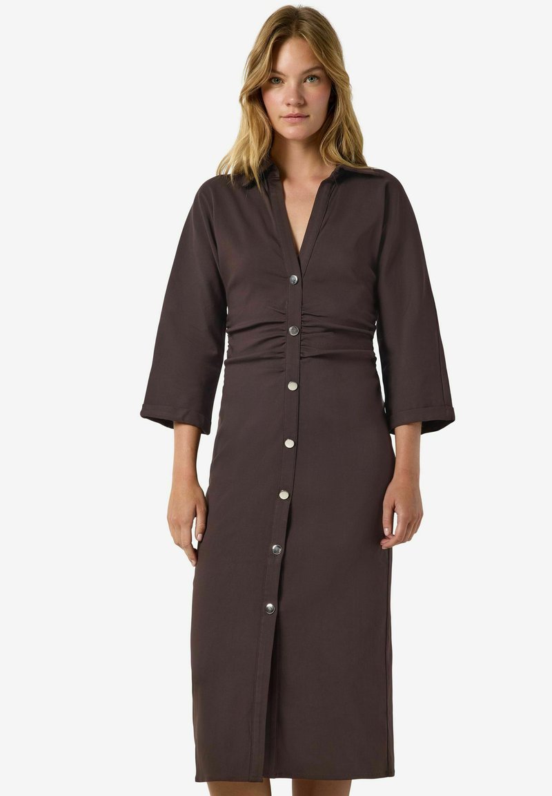 Femme aux cheveux blonds portant une longue robe marron foncé à manches trois-quarts et des boutons argentés sur le devant, debout devant un fond blanc.